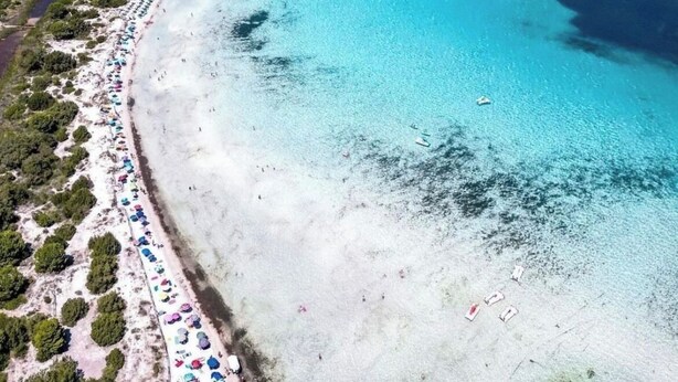 An aerial view of a beach with clear turquoise waters, a long stretch of sandy beach, and a mountainous backdrop.