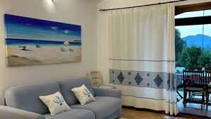 A living room with a grey sofa, a beach-themed painting, a patterned rug, and a view of the outdoors through a window with curtains.