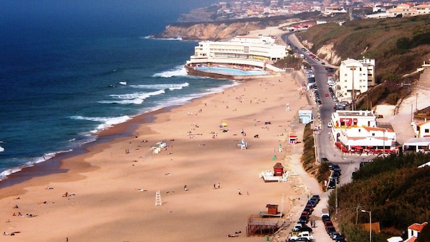 Una playa costera con una carretera que recorre la orilla, un edificio con piscina y un acantilado rocoso.