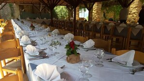 Een lange eettafel gedekt met witte tafelkleden, borden, bestek en wijnglazen, omringd door houten stoelen in een openluchtsetting met stenen muren en begroeiing.