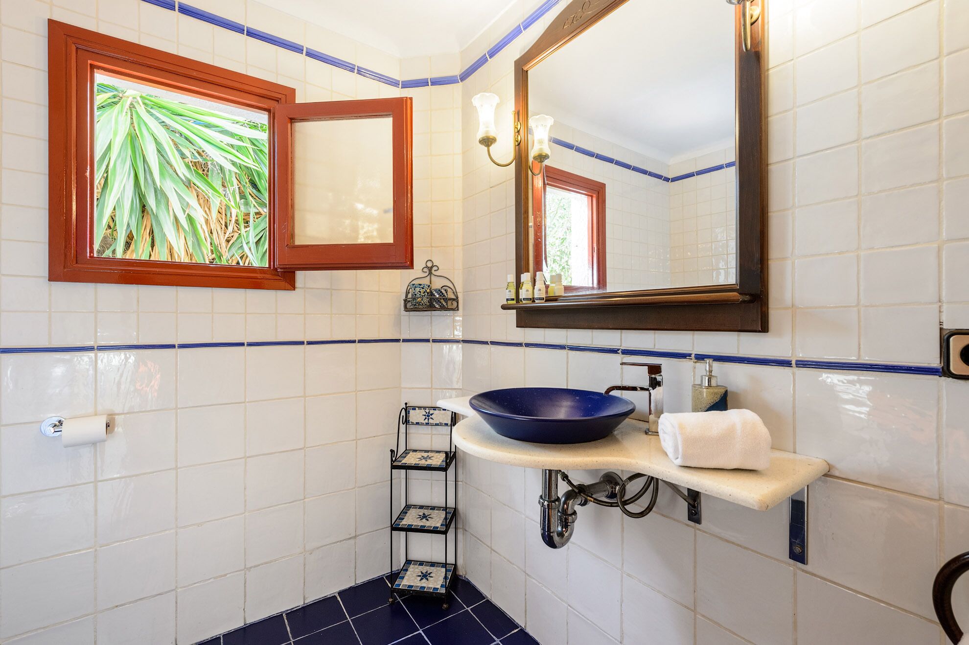 Un baño con paredes de azulejos blancos, suelo de mosaico azul, un espejo grande, un lavabo azul, un perchero y una estantería con artículos de aseo.