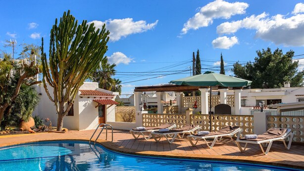 A swimming pool with lounge chairs, a pergola, and a white building with a tiled roof.