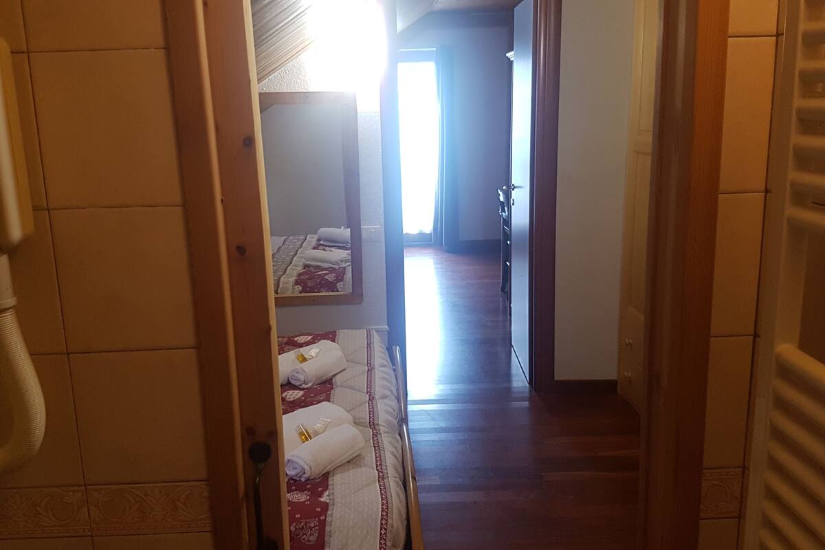 A narrow hallway with wooden flooring, a radiator on the right, and a bed with a red and white patterned bedspread in the background.