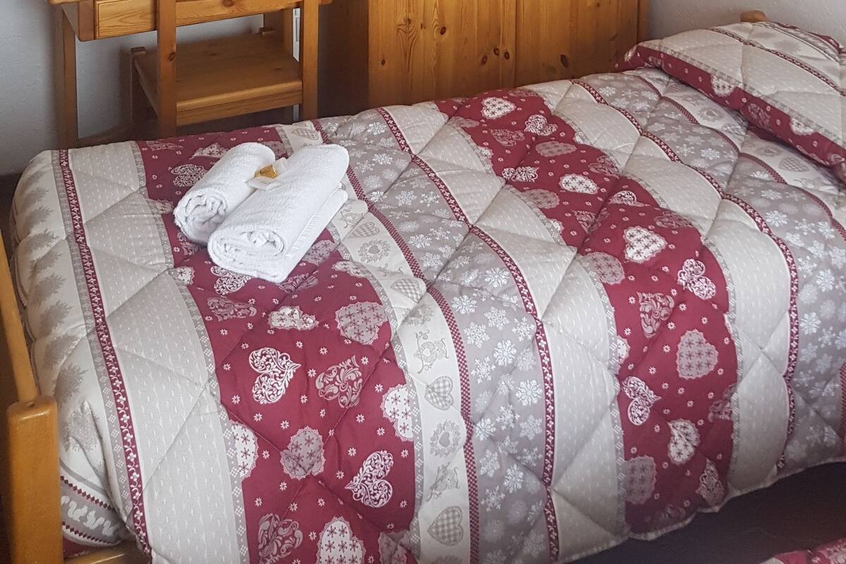 A bedroom with a wooden wardrobe, a wooden desk, a bed with a patterned quilt, and a mirror on the wall.