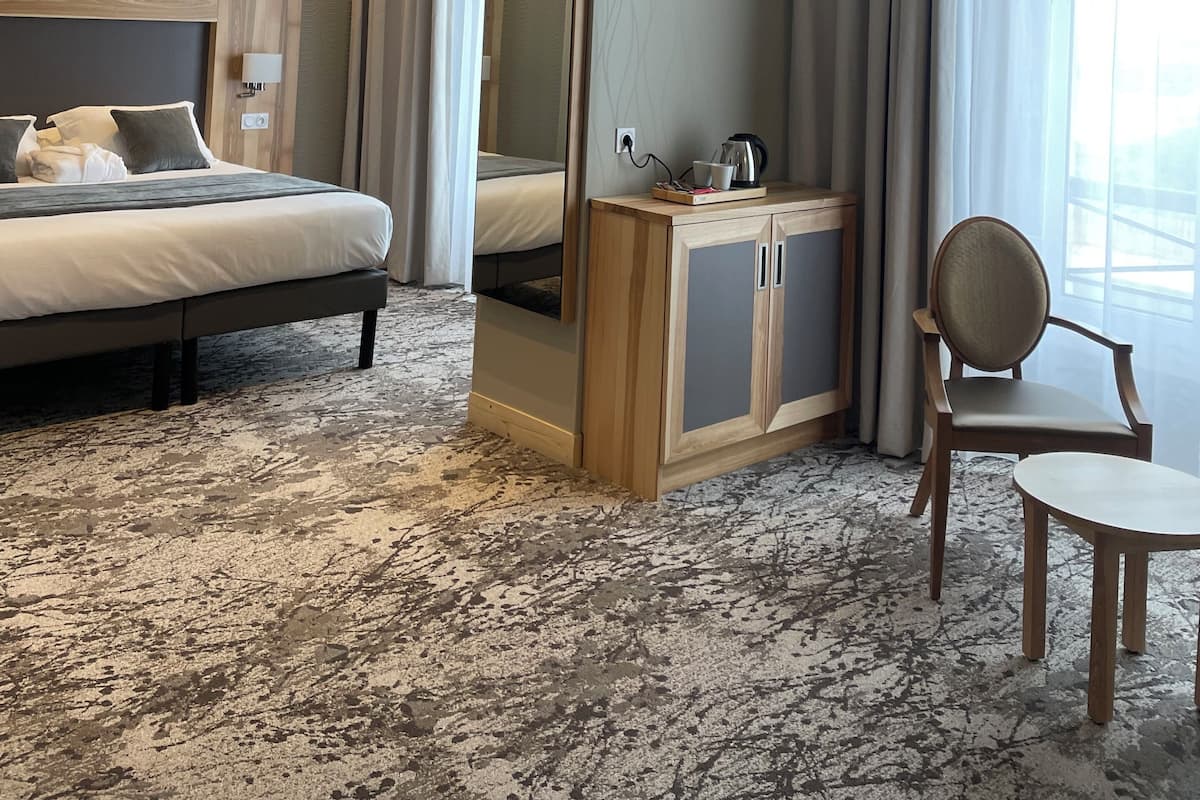 Quarto de hotel com uma cama grande, um armário de madeira, uma cadeira e uma janela ampla com cortinas.