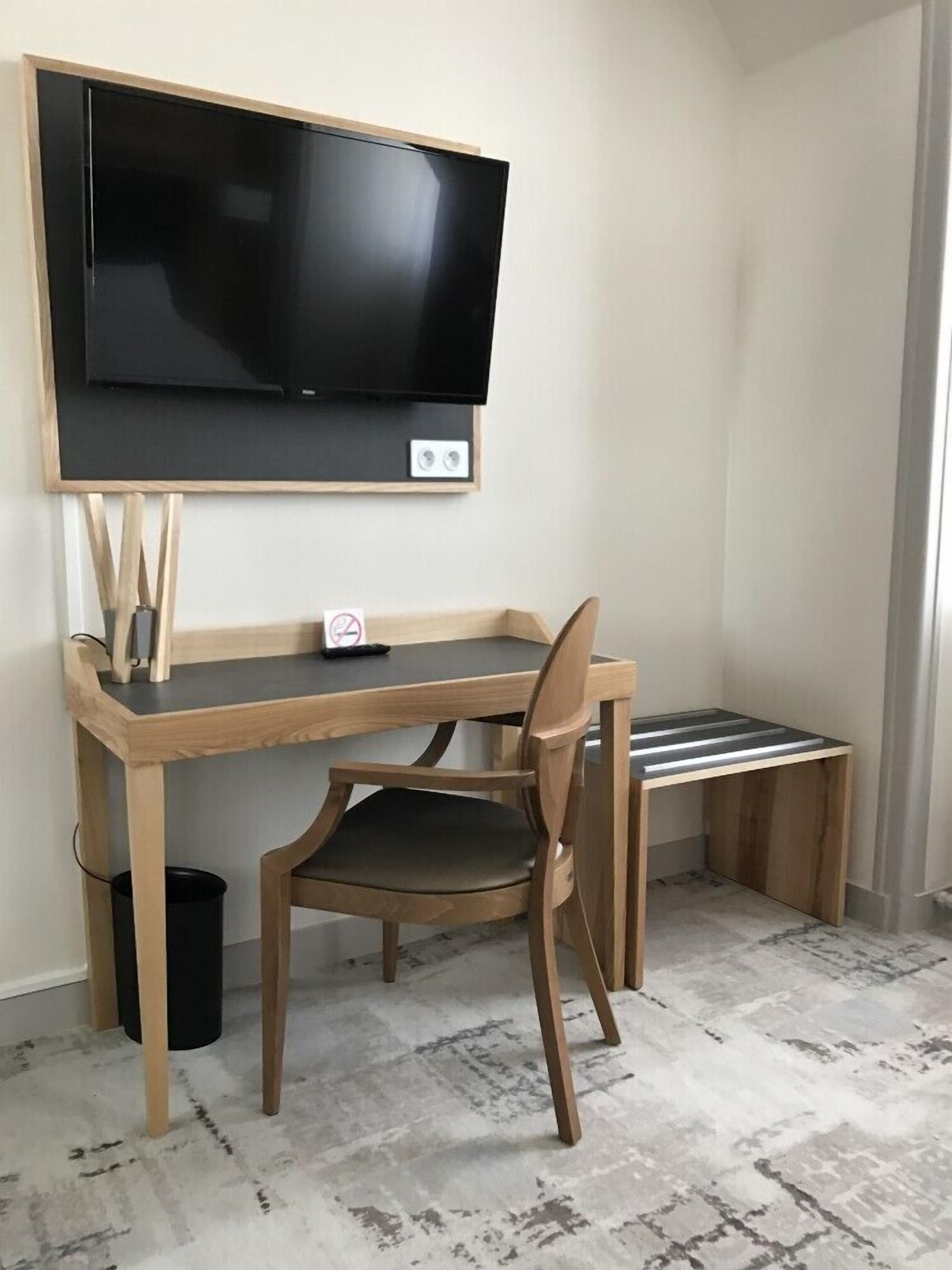 A small, compact room with a wooden desk, a chair, a television mounted on the wall, and a small trash bin.