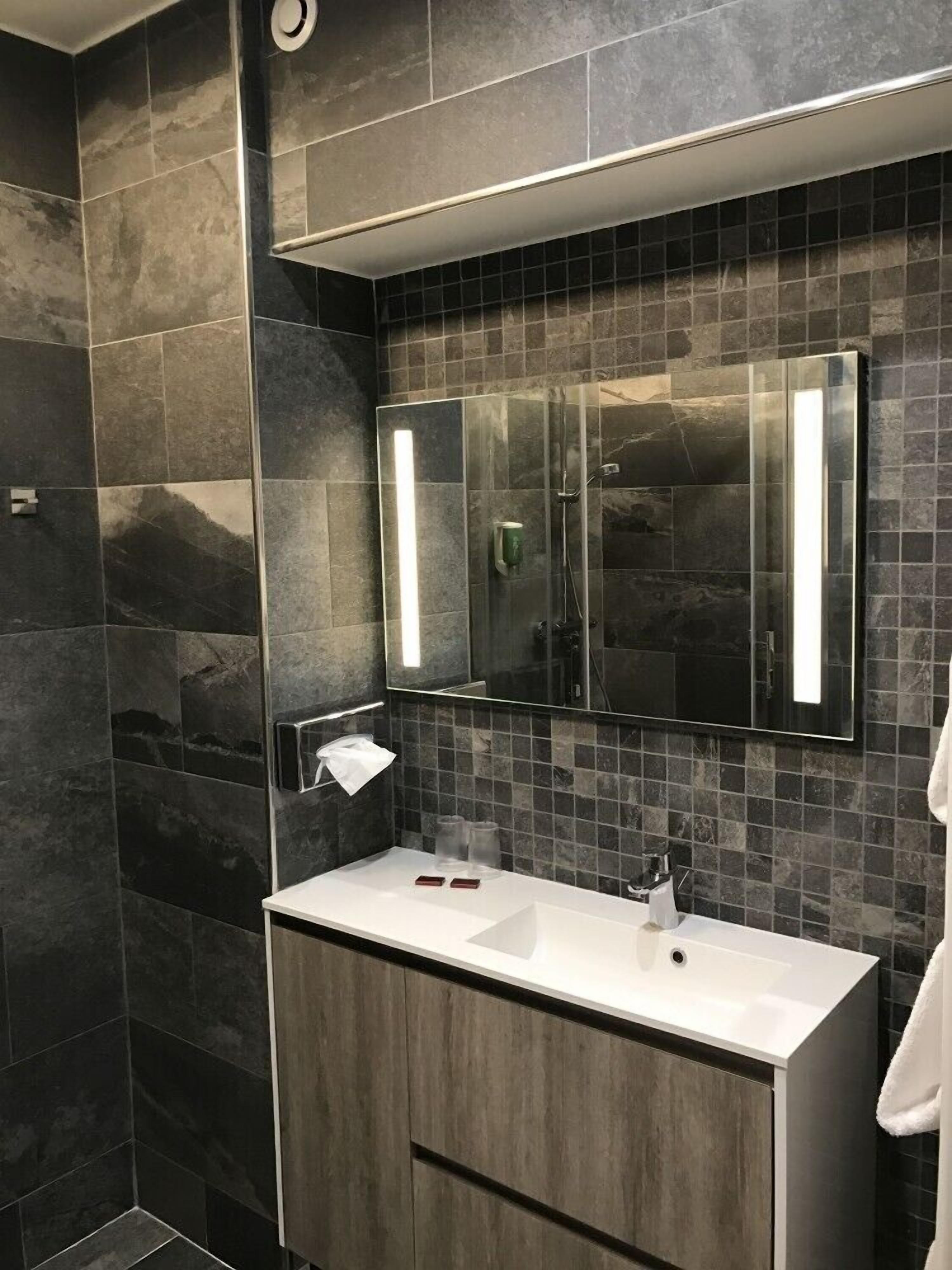 A modern bathroom with dark stone tiles, a large mirror, and a white vanity with a sink.
