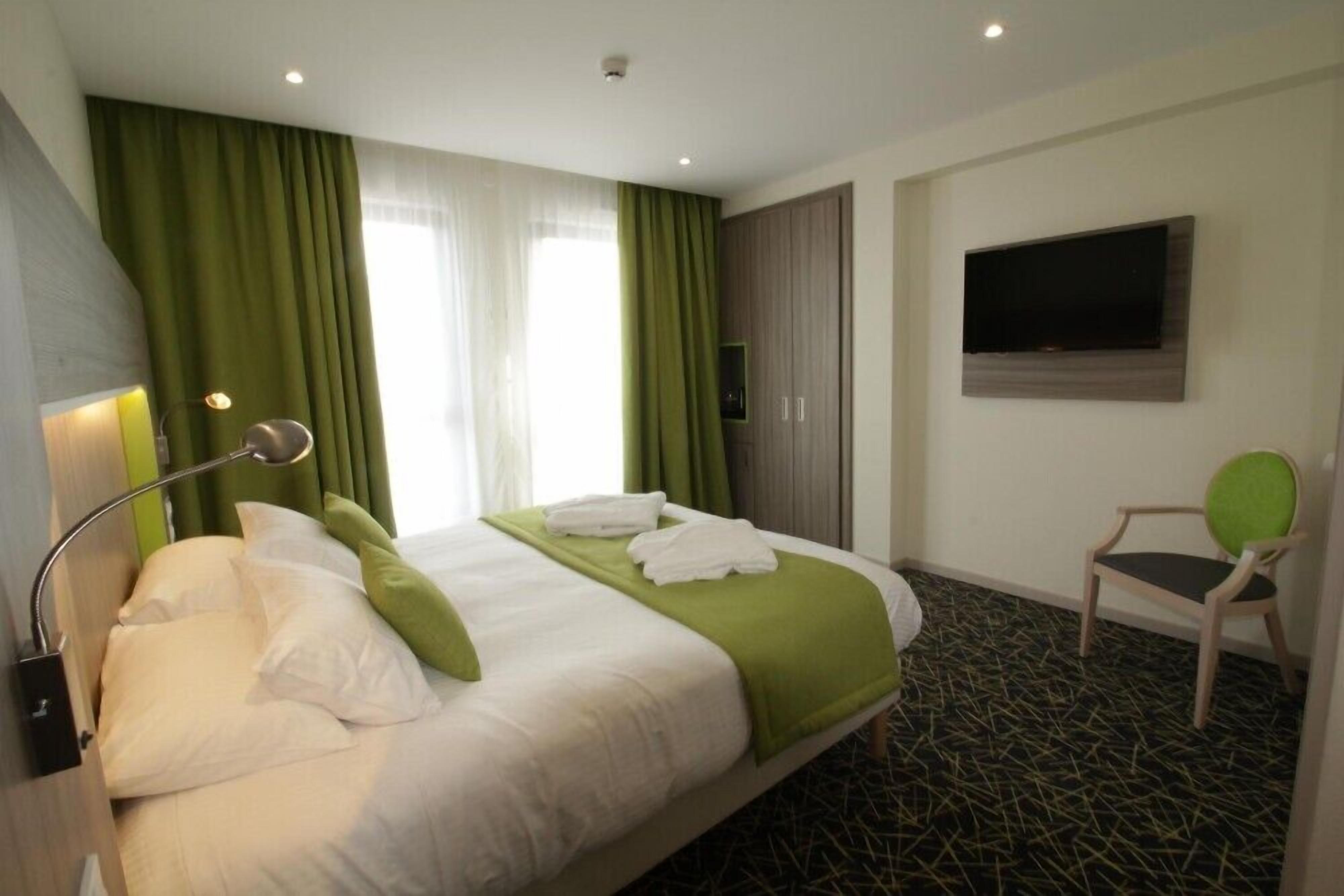 A hotel room with a bed, green curtains, a TV mounted on the wall, and a chair.