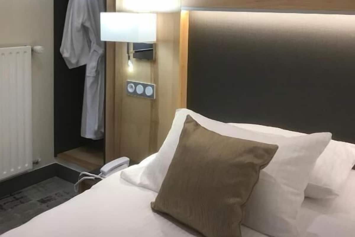 Quarto de hotel com cama, mesa de cabeceira, abajur, guarda-roupa e radiador.