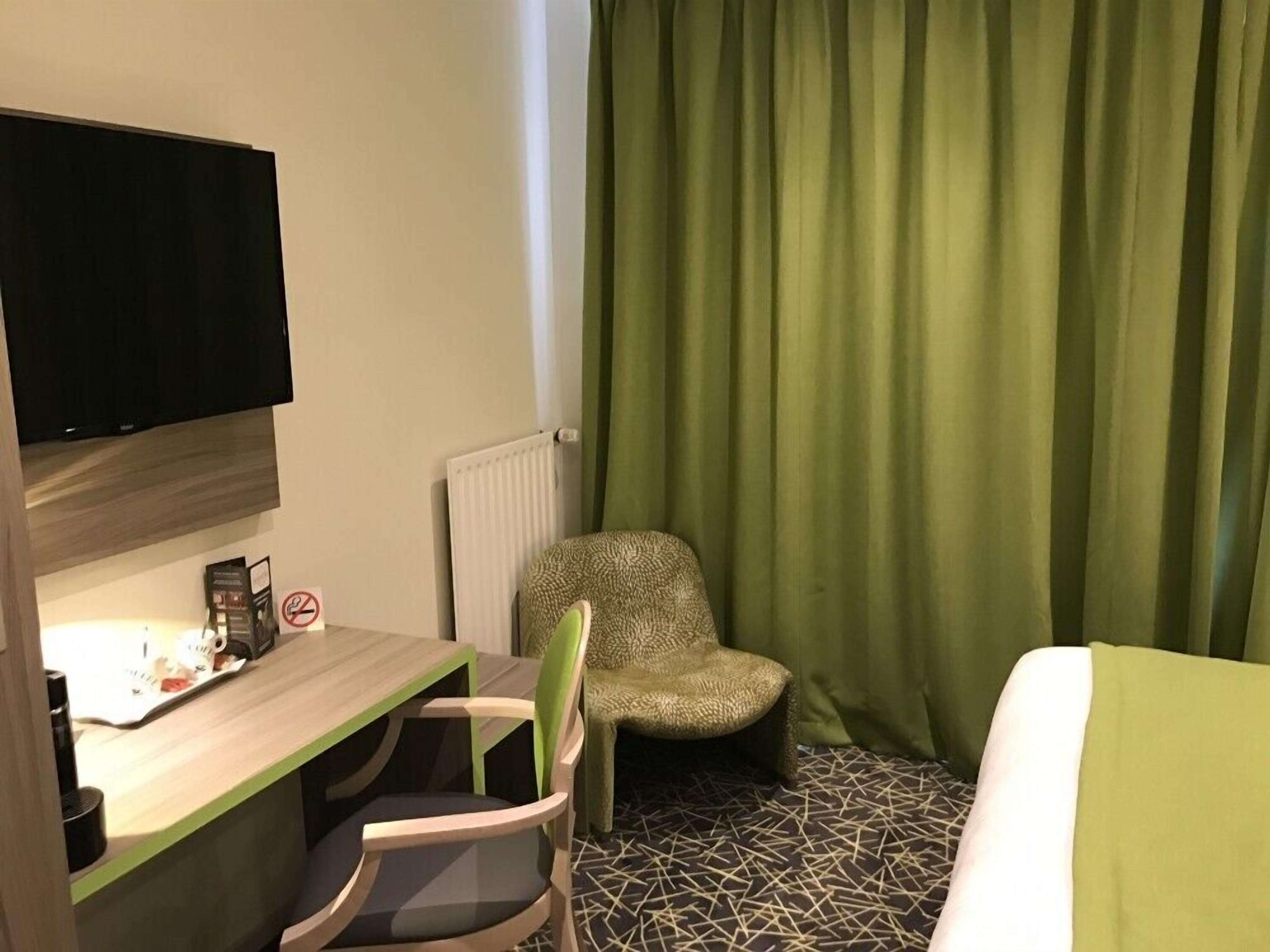 A hotel room with a bed, a desk with a chair, a television mounted on the wall, and a window with green curtains.