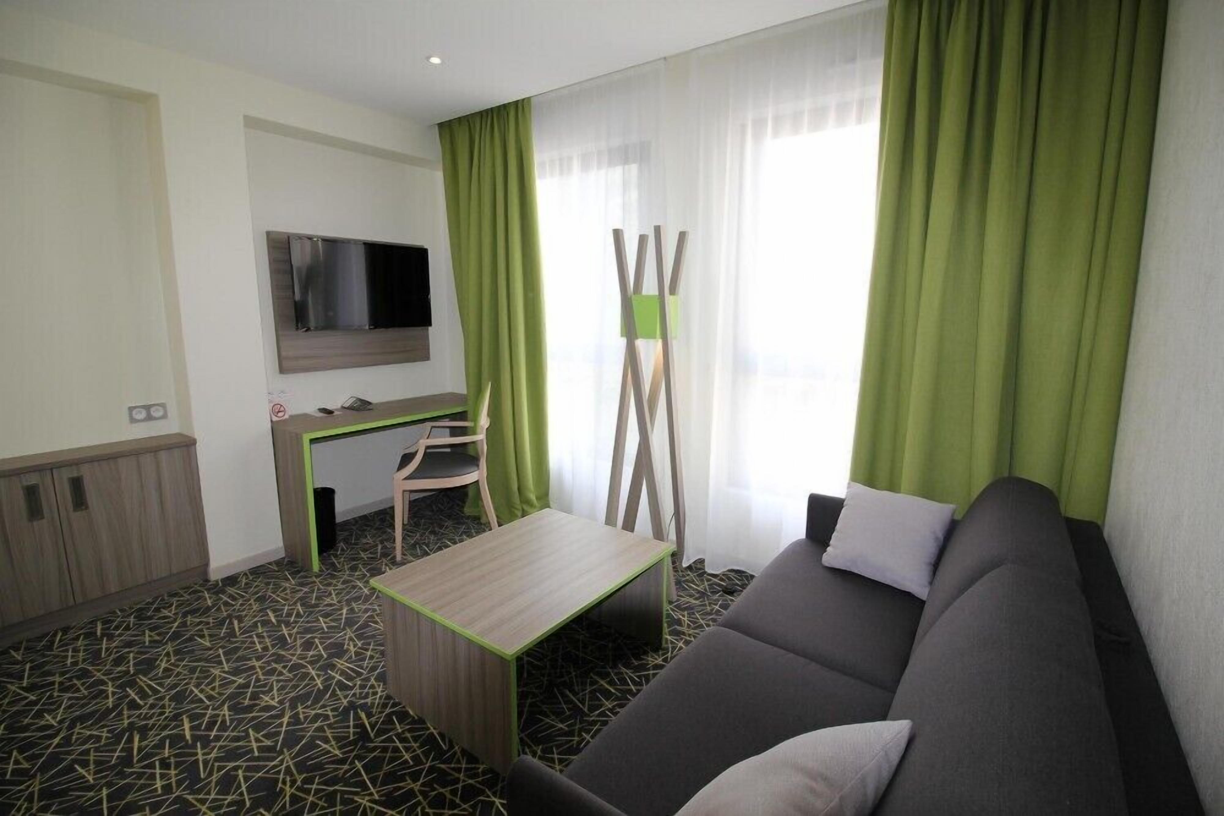 A modern hotel room with a sofa, a desk, a TV mounted on the wall, and green curtains.