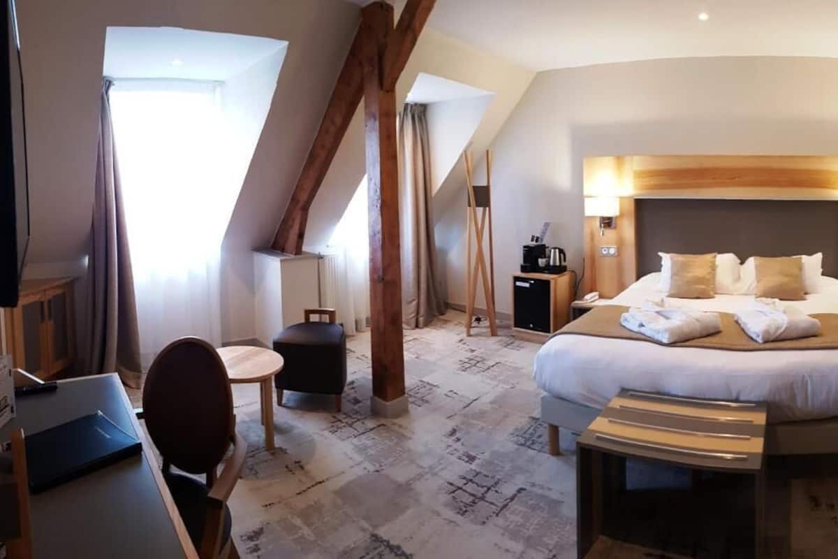 Quarto de hotel com uma cama grande, uma escrivaninha, uma cadeira e uma mesinha de apoio. Há uma janela com cortinas, um abajur e uma máquina de café na mesinha de cabeceira.