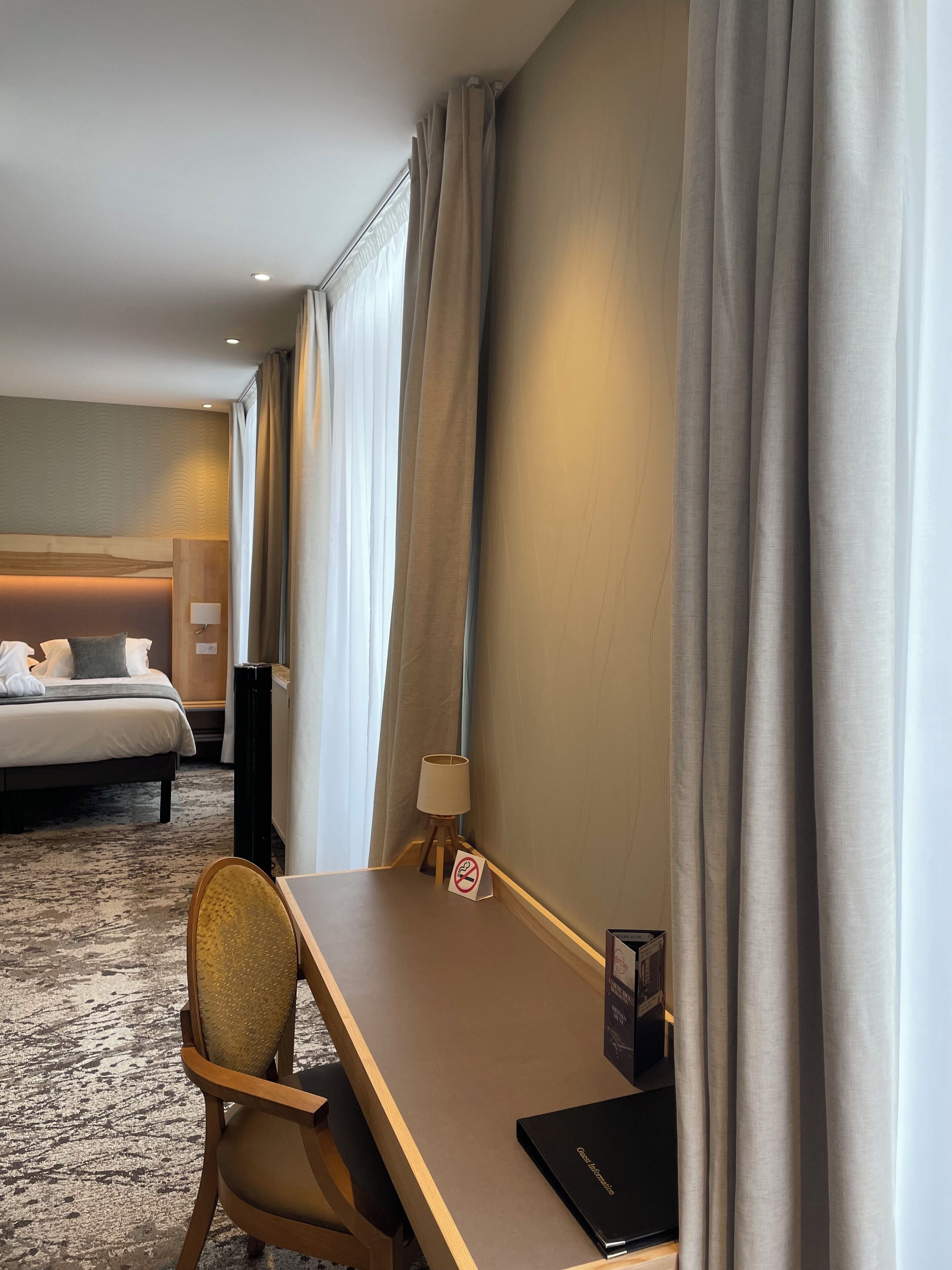 Una habitación de hotel con cama, escritorio, silla, lámpara y cortinas.