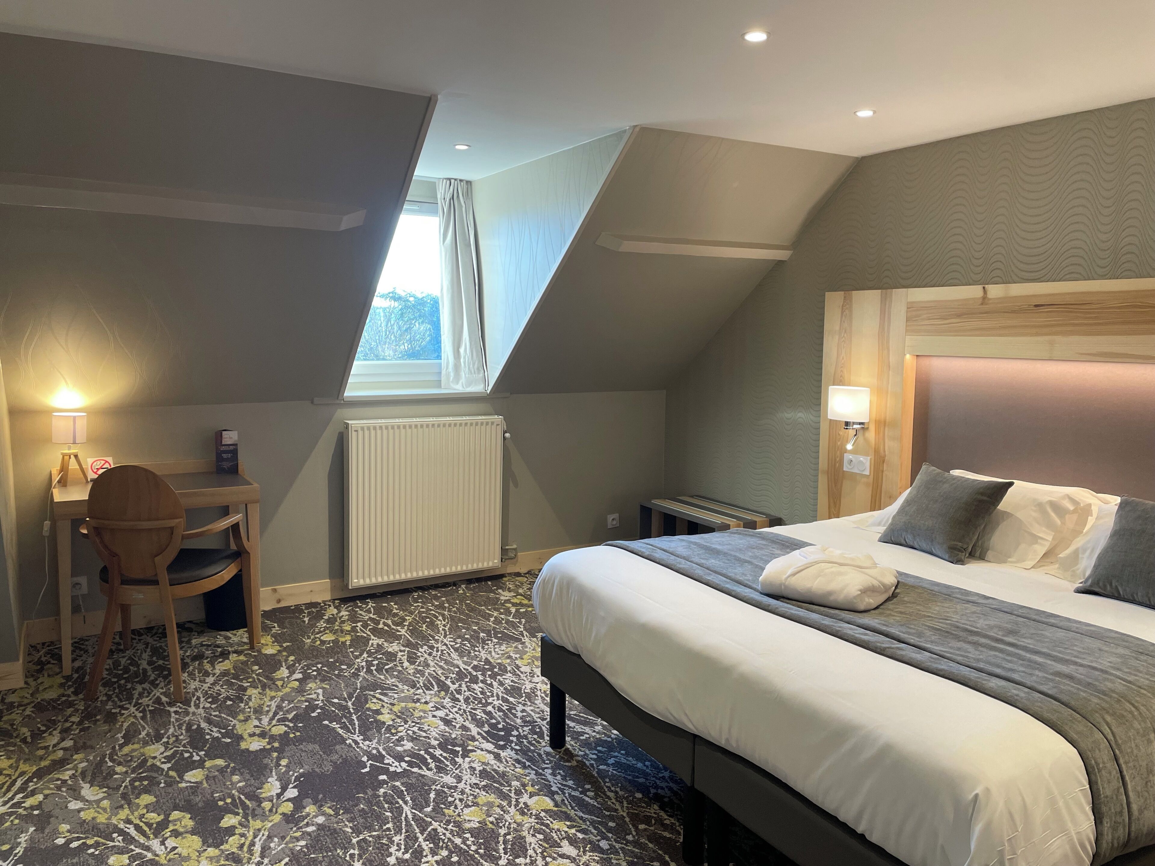 Une chambre d’hôtel avec un lit, un bureau, une chaise, un radiateur et une fenêtre avec des rideaux.