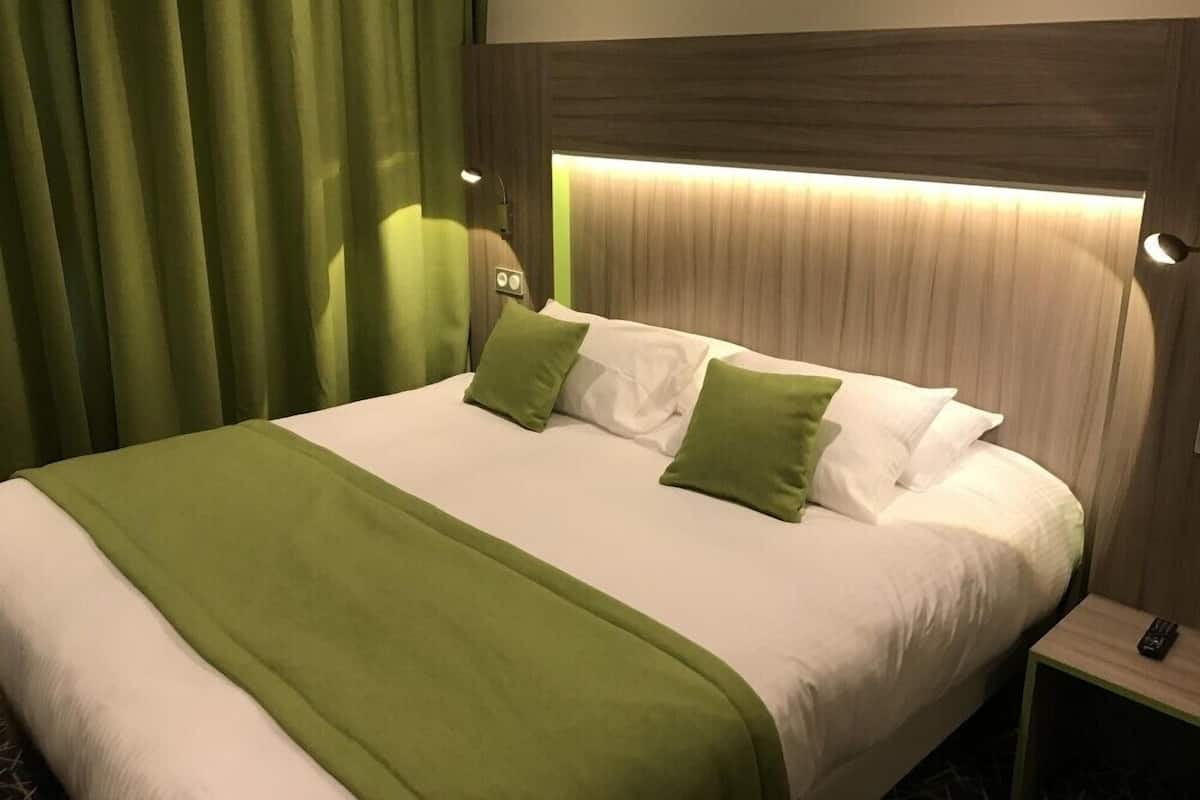 Quarto de hotel com uma cama, cortinas verdes e uma mesa de cabeceira com um controle remoto.