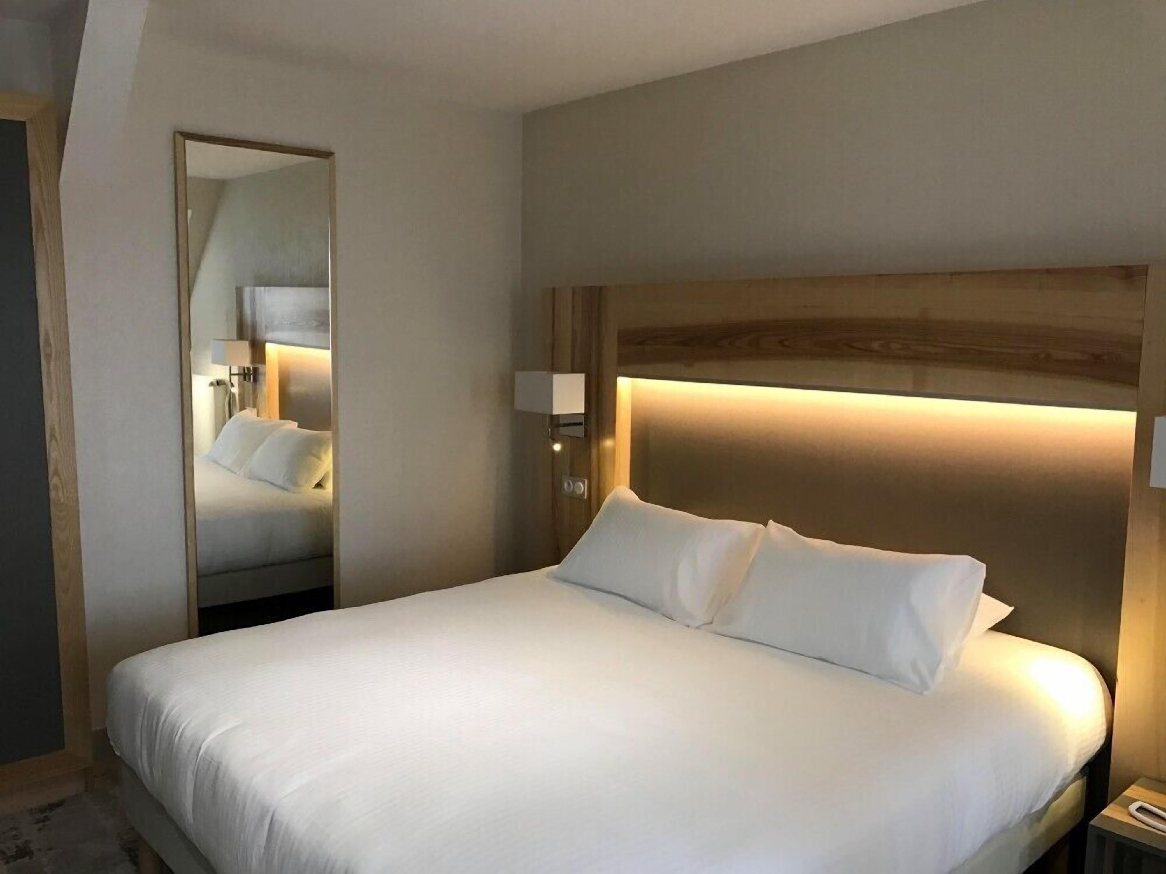 A hotel room with a large bed, a bedside table, a nightstand, a mirror, and a wall-mounted light fixture.
