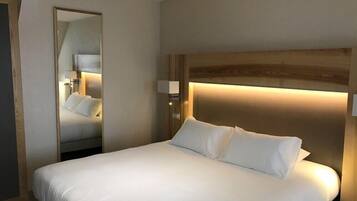 A hotel room with a large bed, a bedside table, a nightstand, a mirror, and a wall-mounted light fixture.