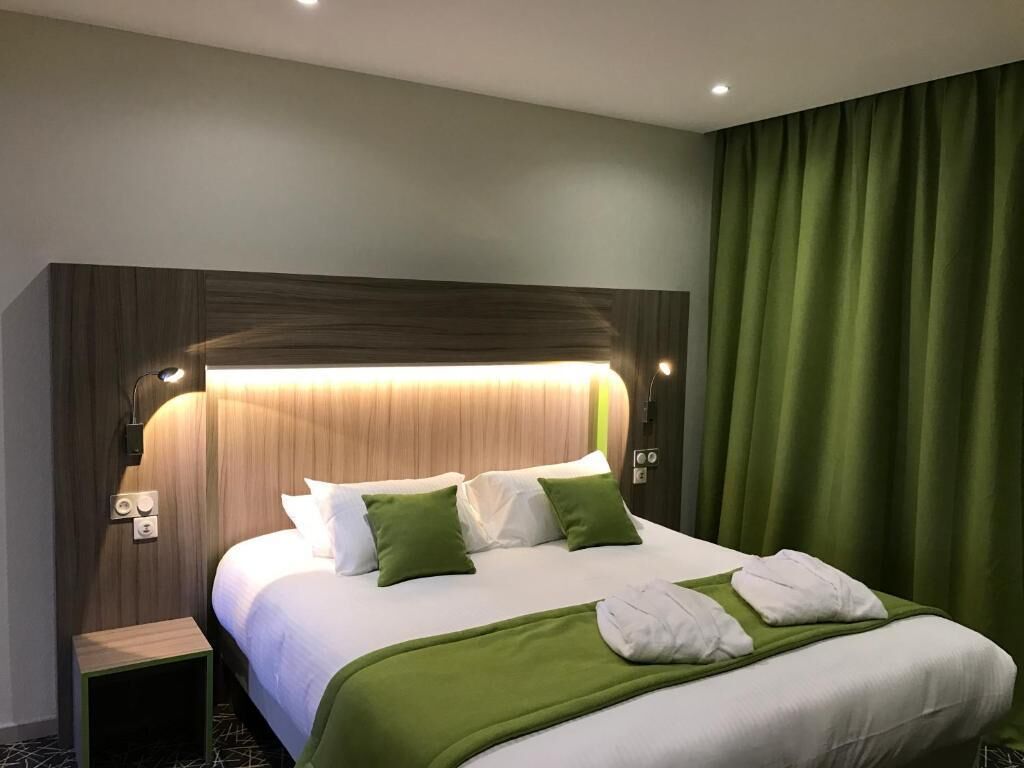 A hotel room with a large bed, green bedding, a headboard with integrated lighting, a nightstand, and a green curtain.