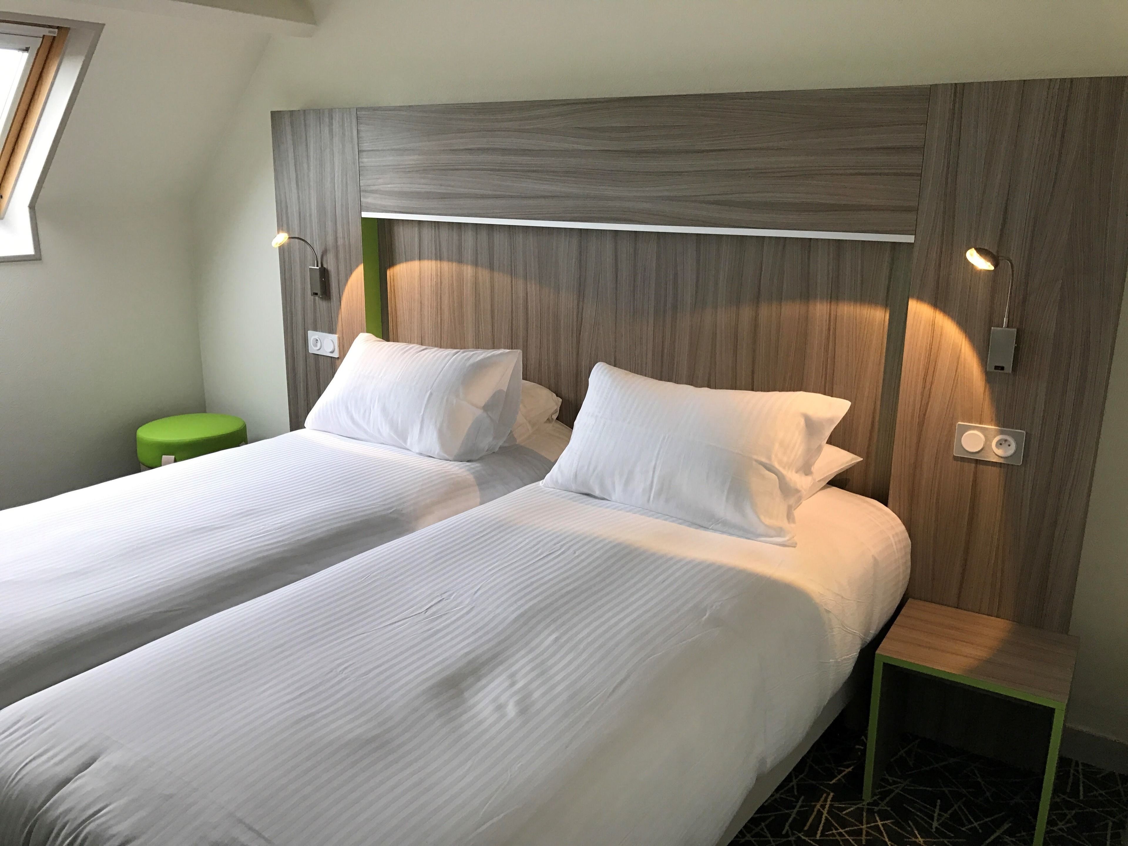 Ett hotellrum med två separata sängar, en grön pall och en sänggavel i trä.