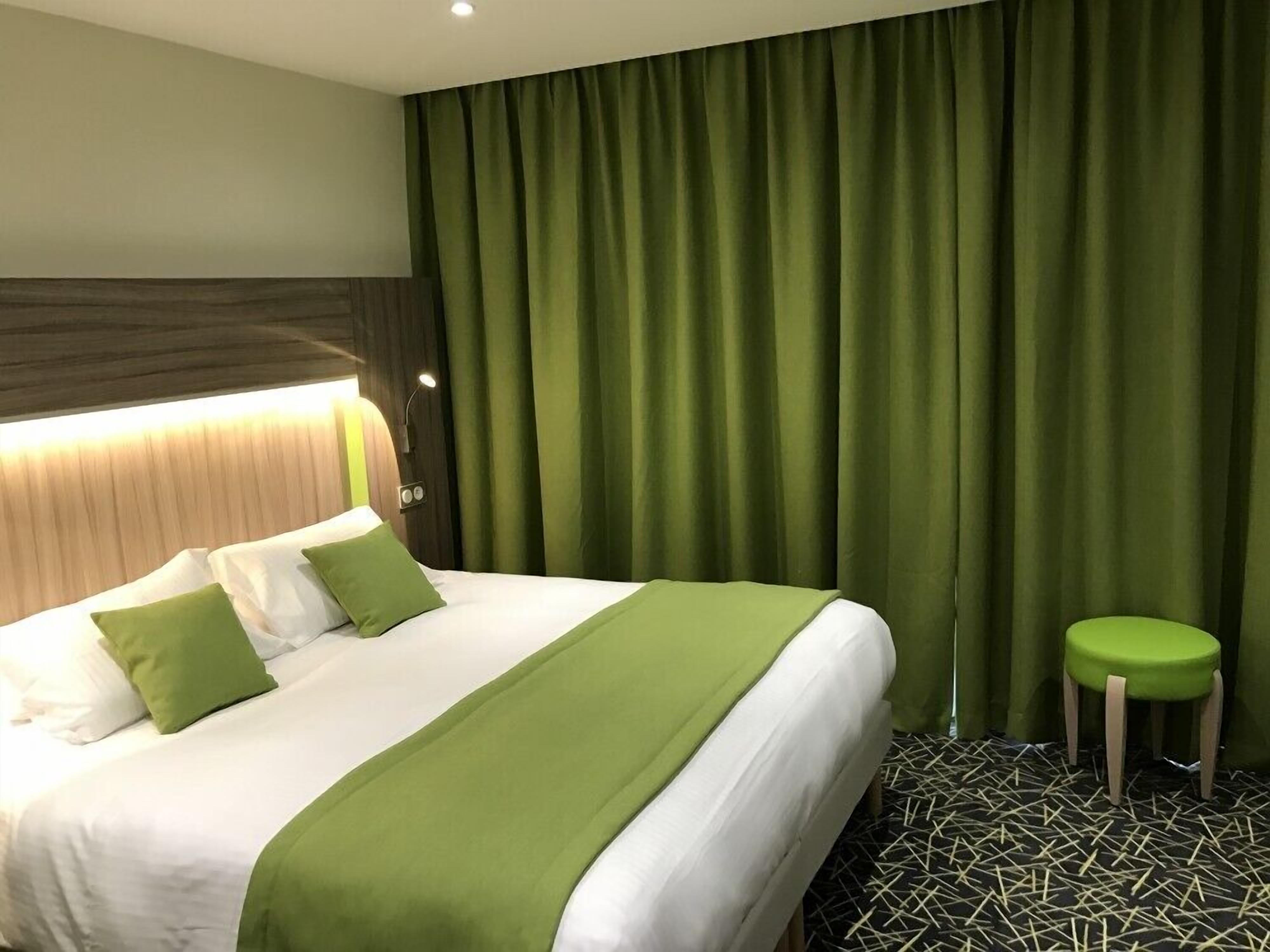 A hotel room with a bed, green curtains, a small stool, and a patterned floor.