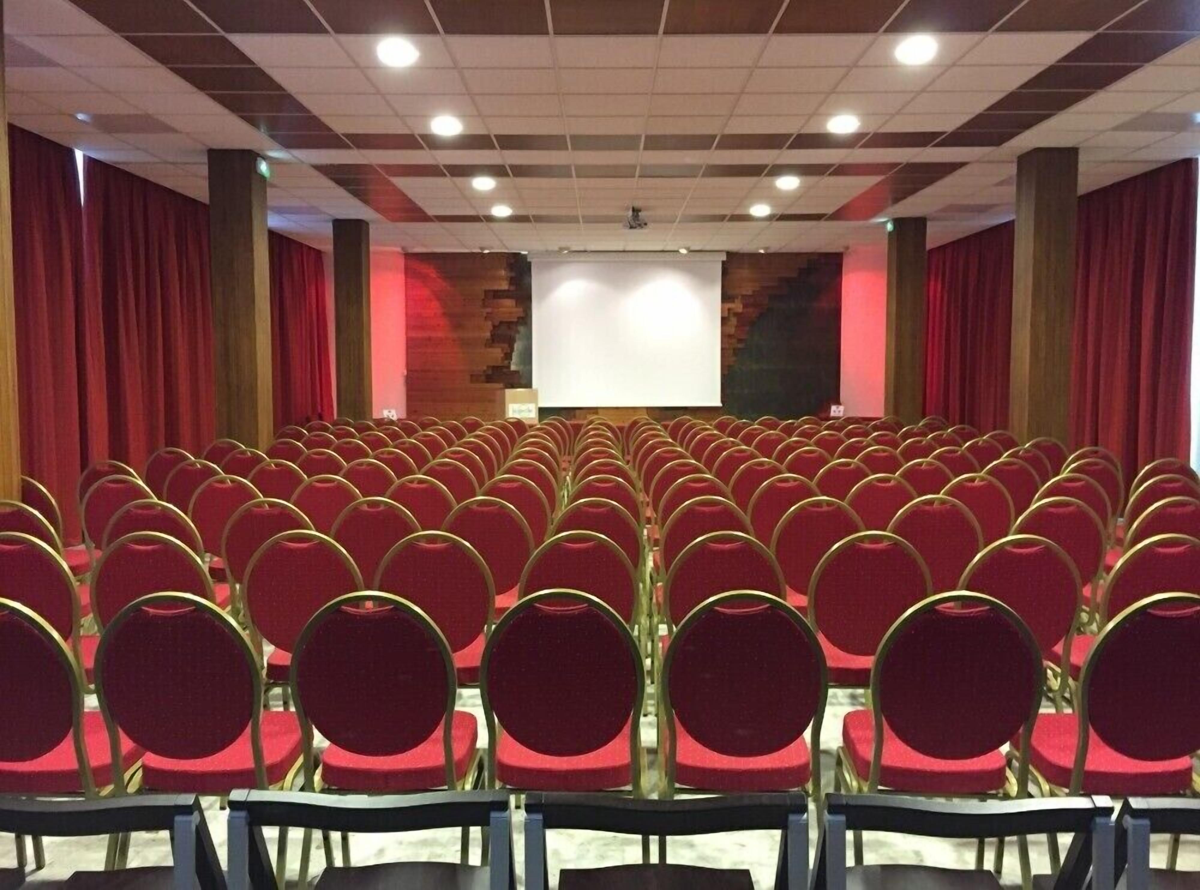 Une salle de conférence avec des rangées de chaises rouges et un grand écran à l’avant.