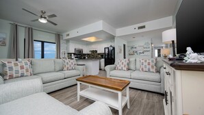 Amplio salón con una mesa de centro de madera, sofás grises con cojines estampados, y una cocina al fondo.