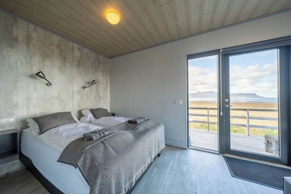 A bedroom with a bed, a concrete wall, a wooden ceiling, and a large window with a view of the beach. - Dis Cottages (Grundarfjörður)