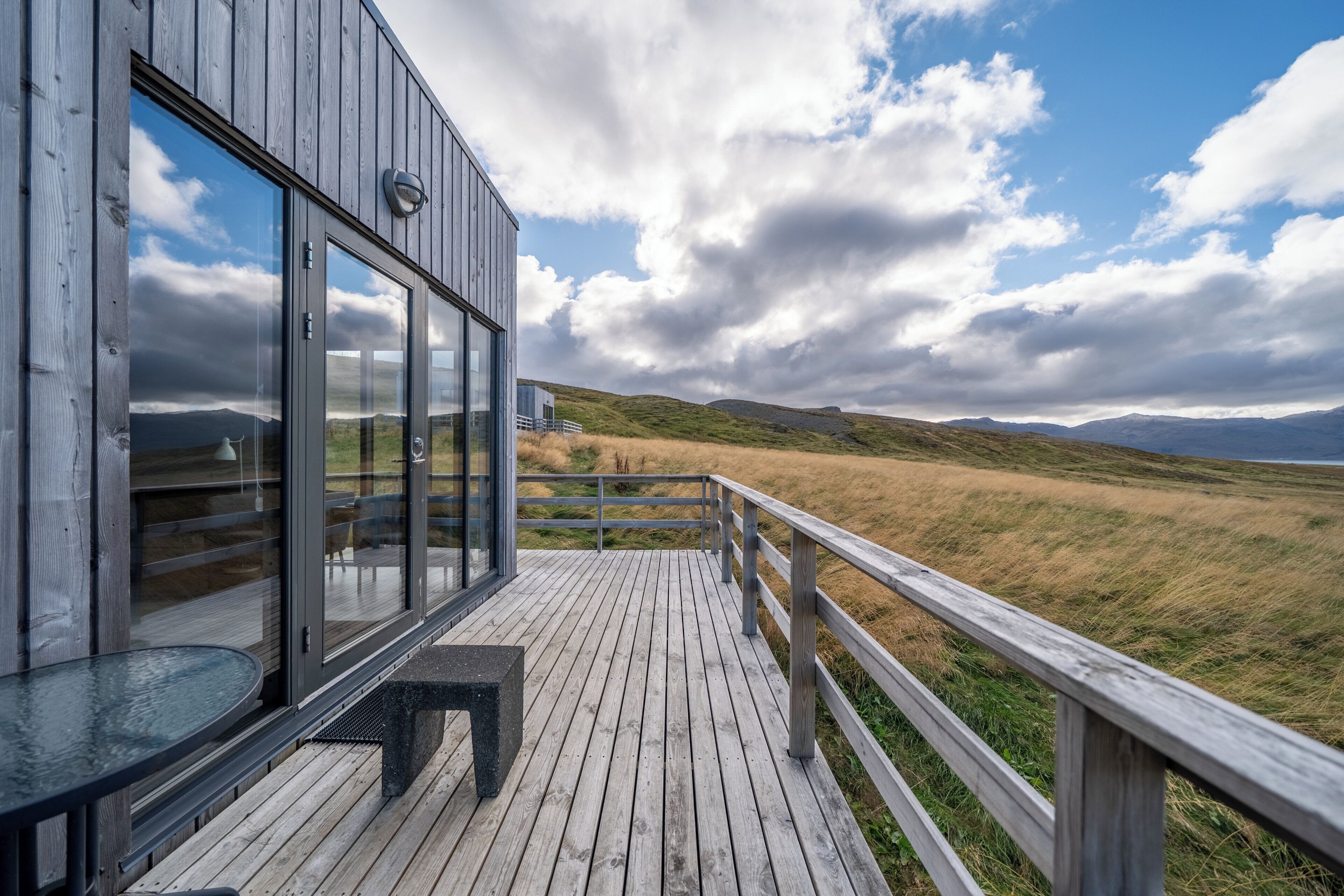 Een houten terras met glazen deuren die uitkomen op een kamer, met uitzicht op een grasachtig landschap en verre bergen.