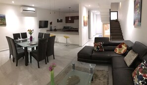 A modern living room with a glass dining table, dark sofas, and a kitchen in the background. - Casa PlayaDelCarmen 515 by Sinbad (Playa del Carmen)