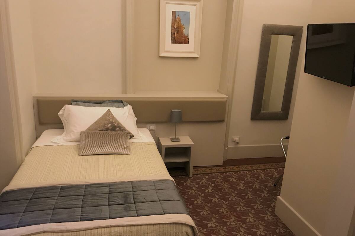 Una habitación de hotel ordenada con una cama, una mesita de noche con lámpara, un cuadro en la pared y un espejo grande.
