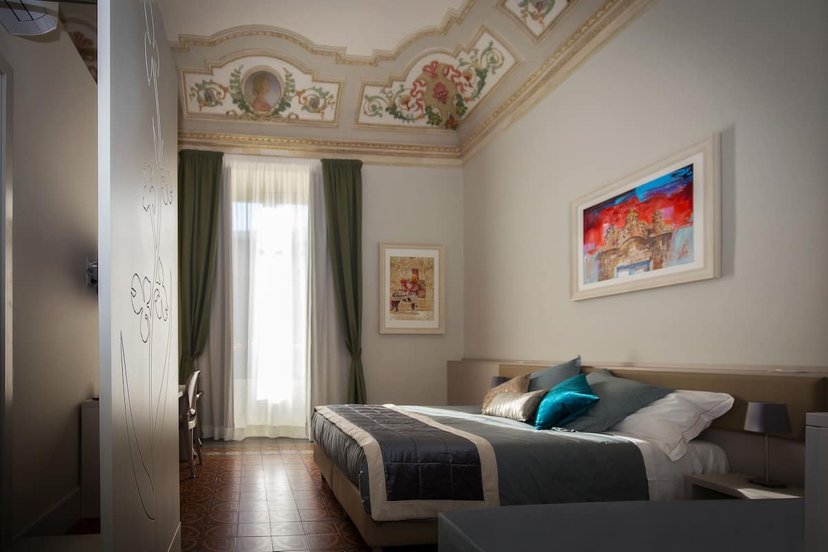 Un dormitorio con cama, mesita de noche, lámpara, un cuadro enmarcado y ventana con cortinas.