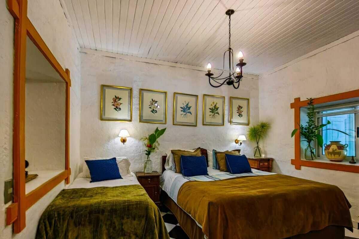 A room with two beds, a checkered floor, a chandelier, framed botanical prints, and a window with plants.
