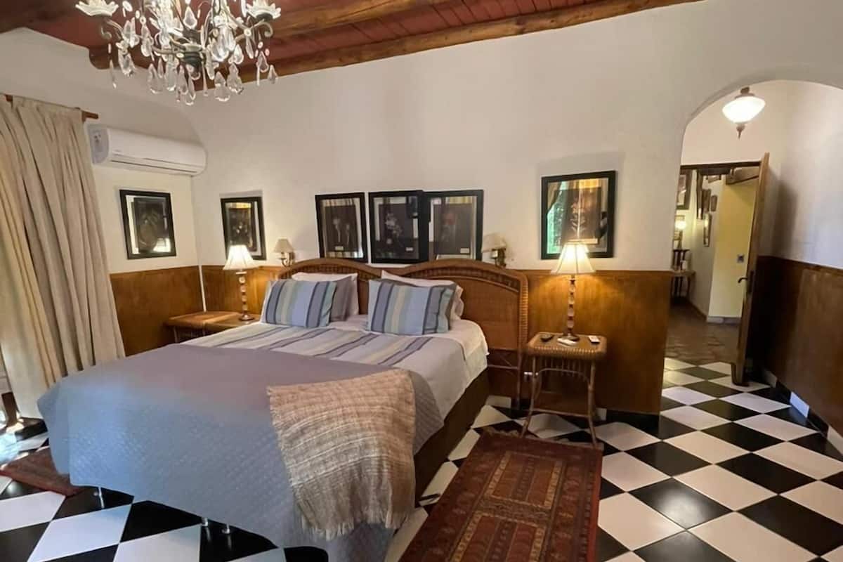 A bedroom with a checkered floor, a bed with a headboard, a chandelier, framed pictures on the wall, and a hallway with a checkered floor.