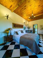 Un dormitorio con suelo a cuadros, techo de madera, cama con manta marrón, mesita de noche con lámpara y aplique de pared.