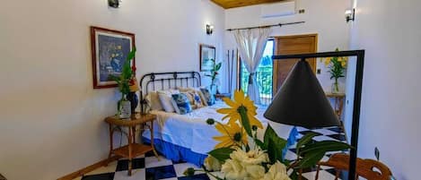A room with a checkered floor, a bed with a white and blue bedspread, a wooden ceiling, and a vase with flowers on a table.