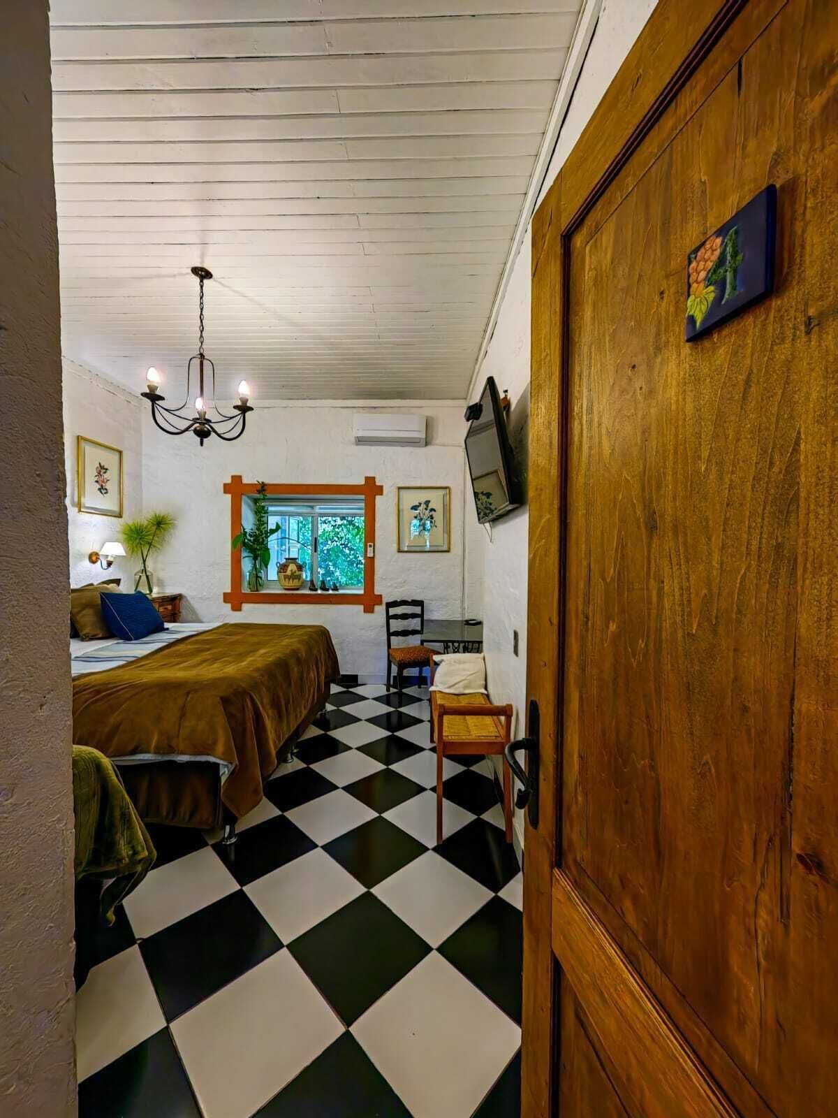 A room with a checkered floor, a bed with a brown bedspread, a wooden door, and a window with a view of greenery.
