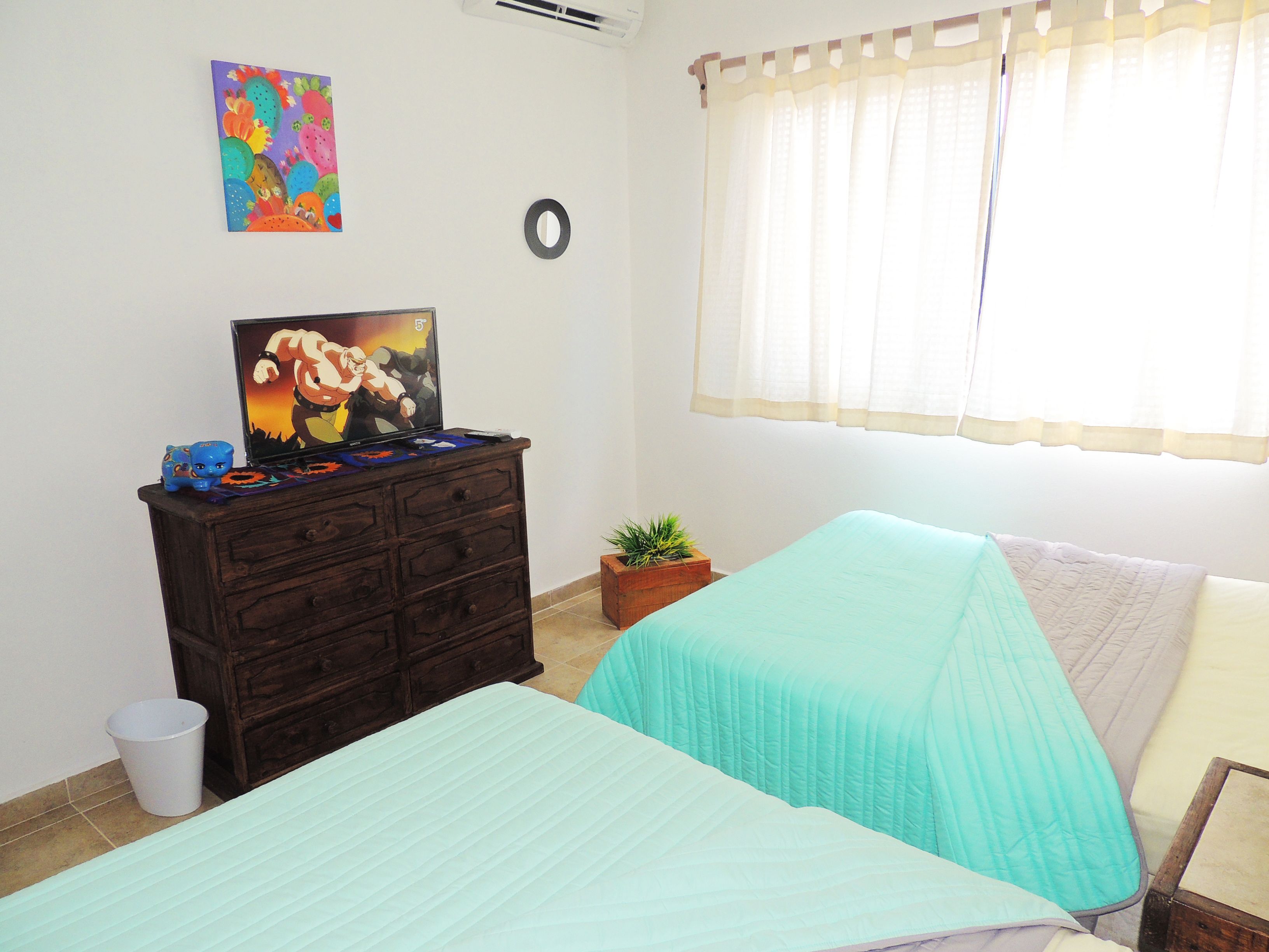 Une chambre avec deux lits, une commode, un tableau coloré, une fenêtre avec des rideaux et une petite plante.