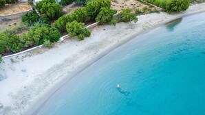 An aerial view of a beach with clear turquoise water, a person in the water, and a row of beach umbrellas along the shore. - Maison Simone, a great escape experience!! (Αγ. Σώστης)