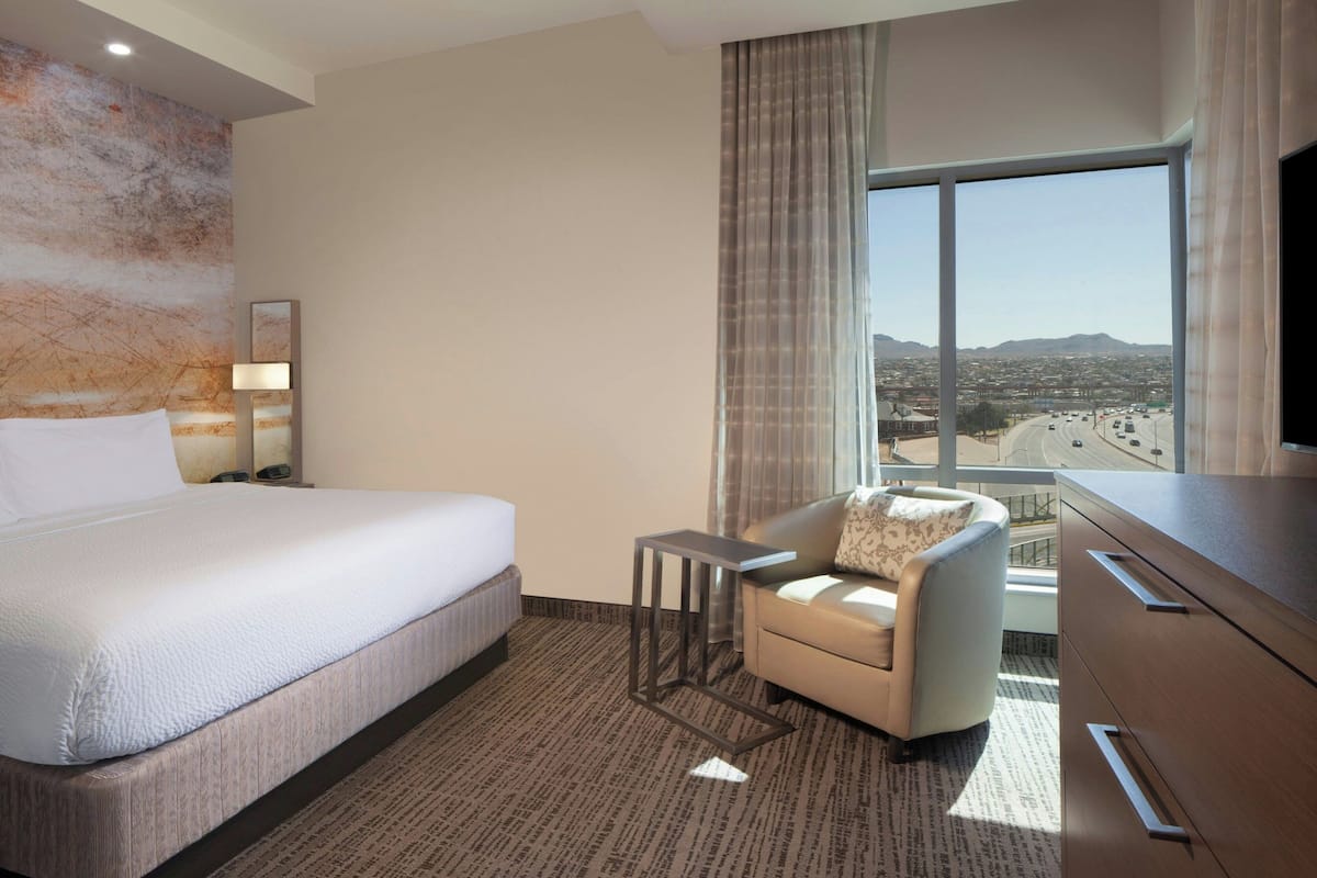 Ein Hotelzimmer mit einem großen Bett, einem kleinen Tisch, einer Sitzecke, einem Fernseher und Blick auf eine Stadtlandschaft durch ein großes Fenster.