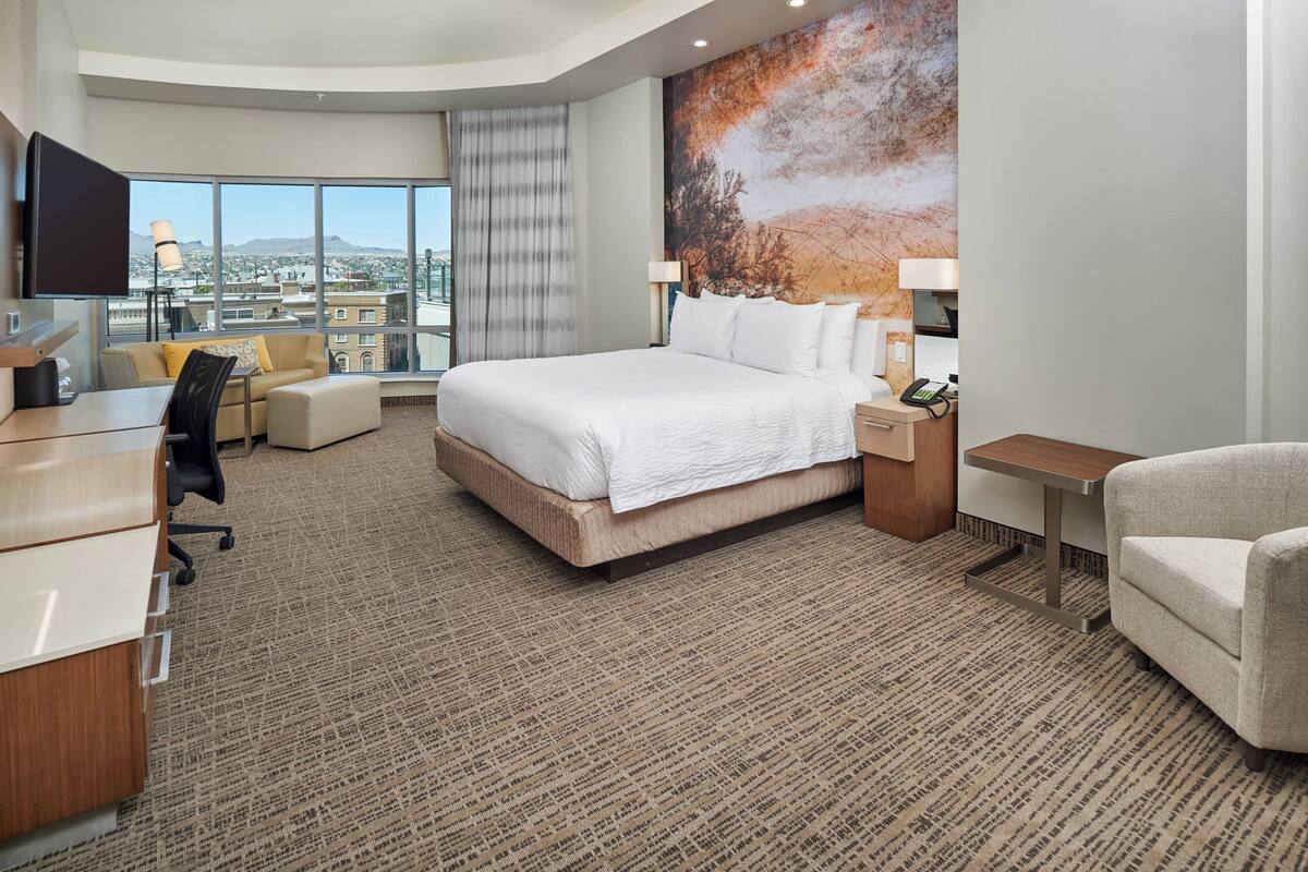 Ein Hotelzimmer mit einem großen Bett, einem Schreibtisch, einem Stuhl und Blick auf die Stadt durch große Fenster.