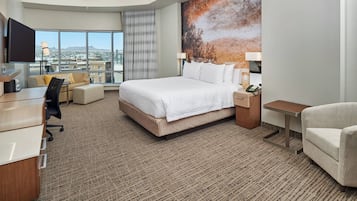 Habitación de hotel con una cama grande, un escritorio, una silla y vistas de la ciudad a través de amplios ventanales.