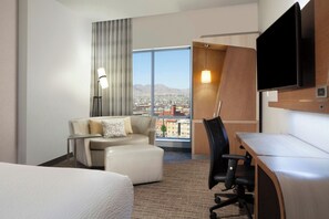 Hotelkamer met een bed, bureau, stoel, bank, televisie, lamp en een groot raam met uitzicht op de stad.