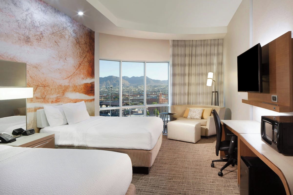 Ein Hotelzimmer mit zwei Betten, einem Schreibtisch mit Fernseher, einer Schreibtischlampe und Blick auf eine Stadtlandschaft.