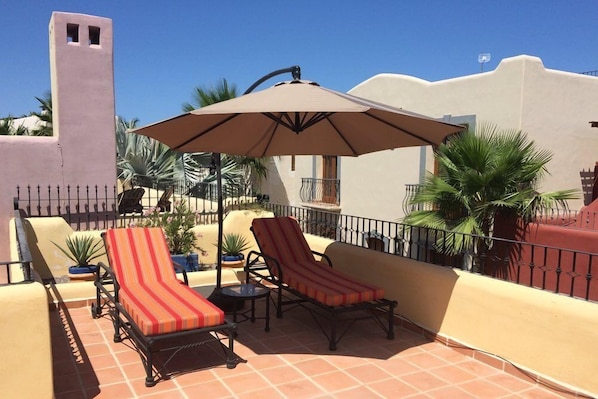 A terrace with two lounge chairs and a table, surrounded by potted plants and a large umbrella.