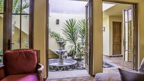 Une pièce avec un fauteuil rouge, un lit avec un oreiller et une vue sur une cour intérieure agrémentée d’une fontaine et de plantes.