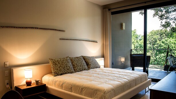 A bedroom with a large bed, a bedside table with a lamp, a chair, and a view of the outdoors through a balcony. - Depto en Cancun 506 by Sinbad (Cancun)