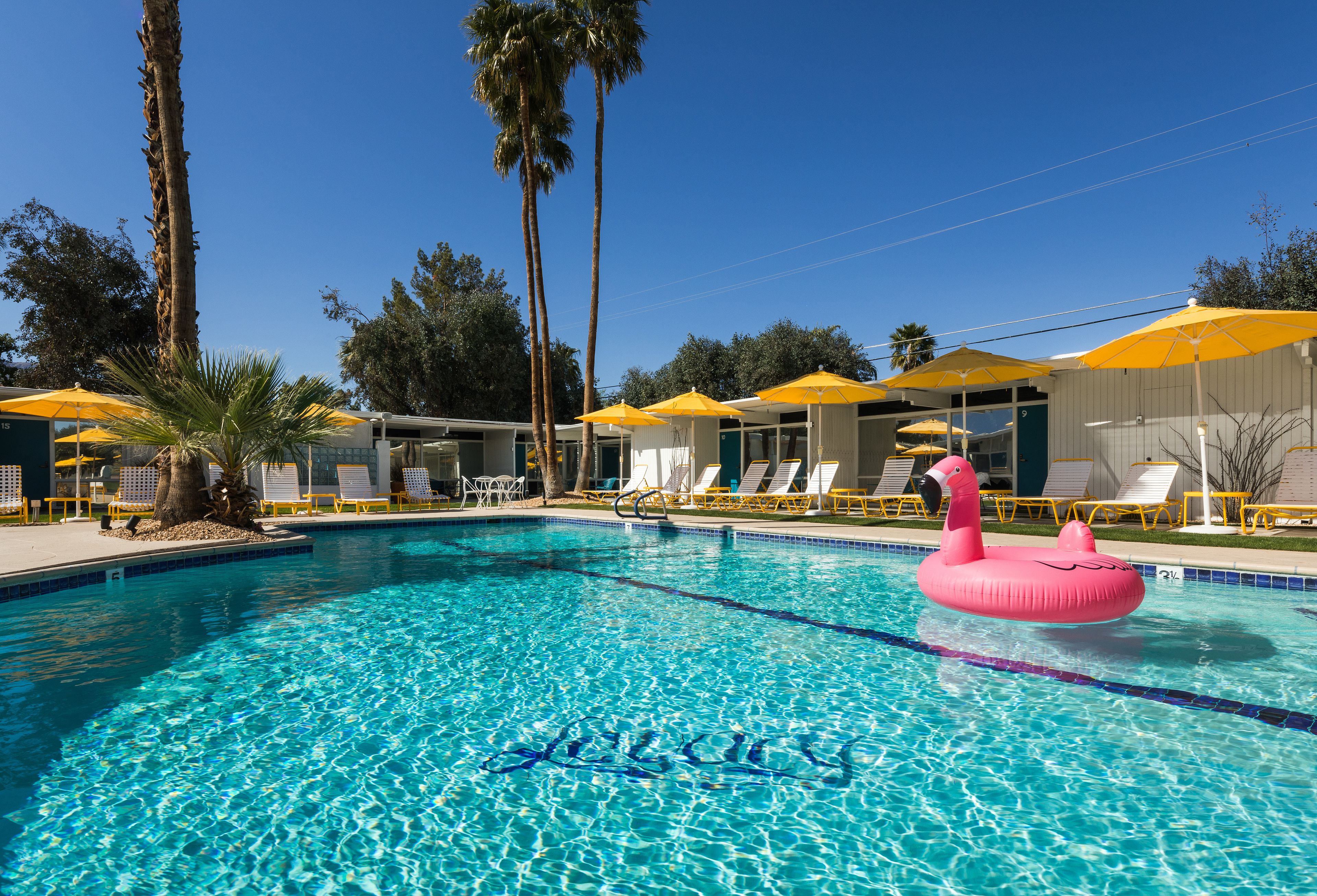 A clear blue pool with a pink flamingo float, surrounded by lounge chairs and yellow umbrellas.