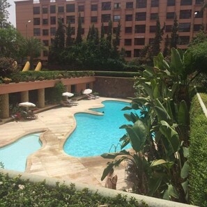 A swimming pool with a wavy edge, surrounded by lounge chairs and umbrellas, with a building in the background.