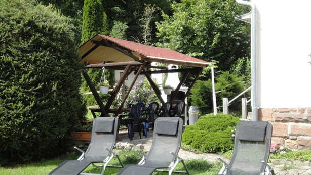 Tres tumbonas en un jardín con una pérgola de madera y un muro de piedra al fondo.