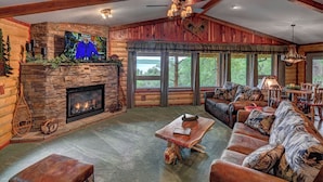 A cozy log cabin interior with a stone fireplace, leather furniture, and large windows offering a view of the outdoors.